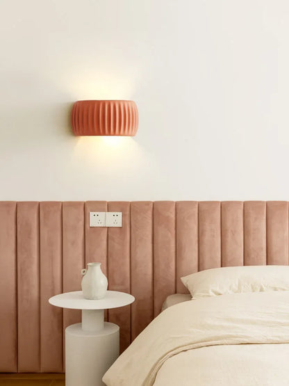 A modern bedroom scene featuring a ribbed terracotta orange wall sconce mounted on a white wall above a dusty rose velvet headboard. A small white side table with a white vase sits next to a bed with cream bedding.